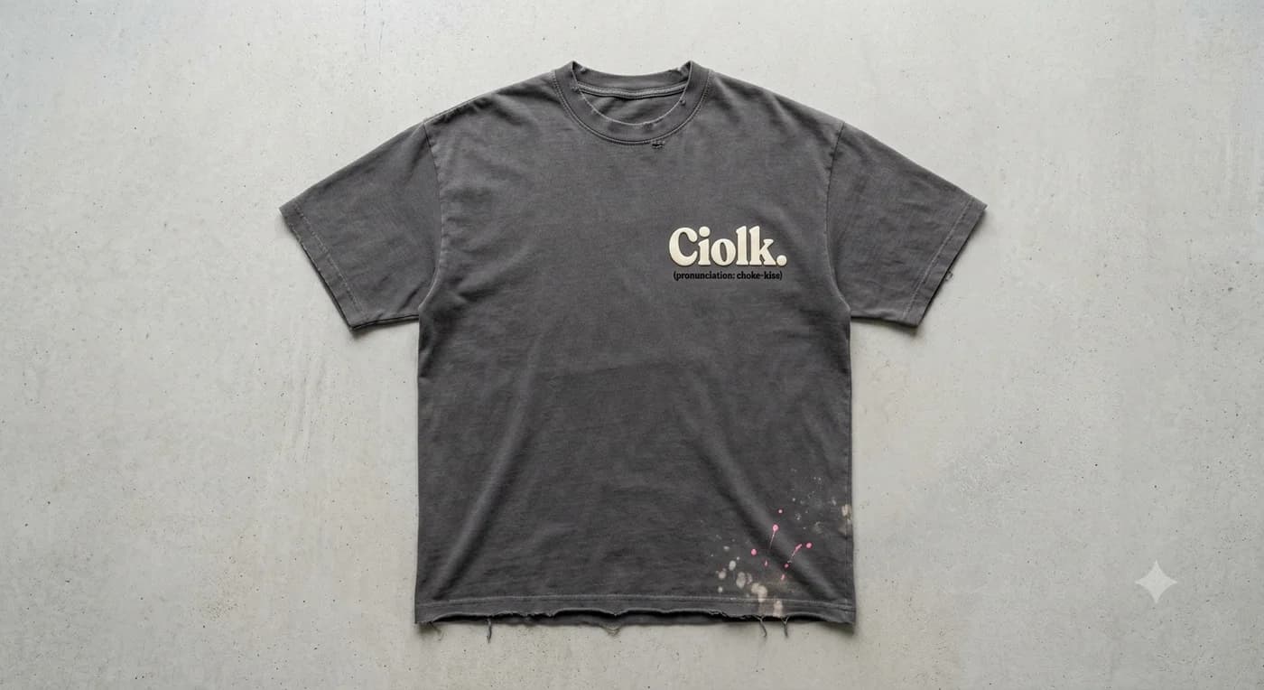 A washed charcoal T-shirt laid flat on concrete, printed on the chest with Ciolk. and the pronunciation choke-kise, with a frayed hem and pink and cream paint flecks near the bottom.